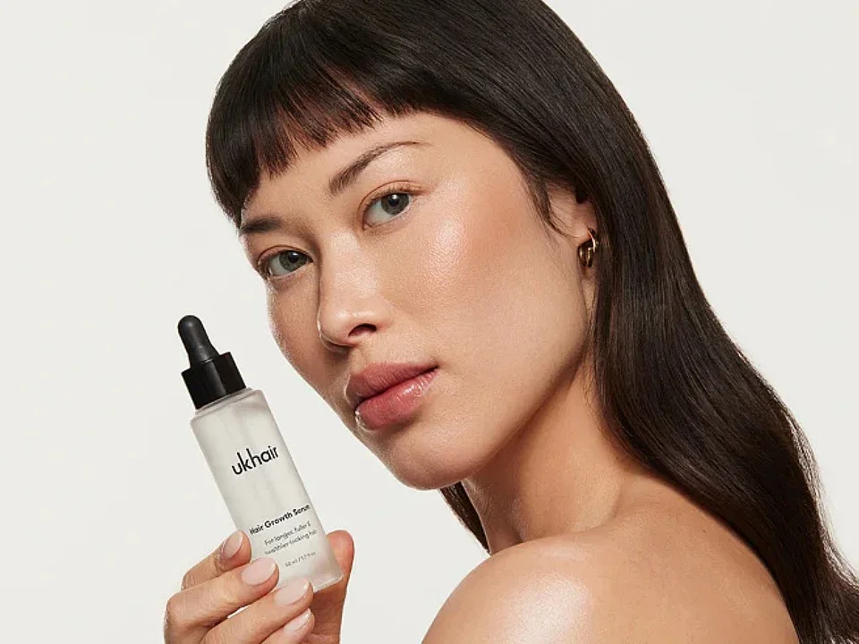 Woman with straight dark hair holding a frosted dropper bottle labeled ‘UKHAIR Hair Growth Serum’.