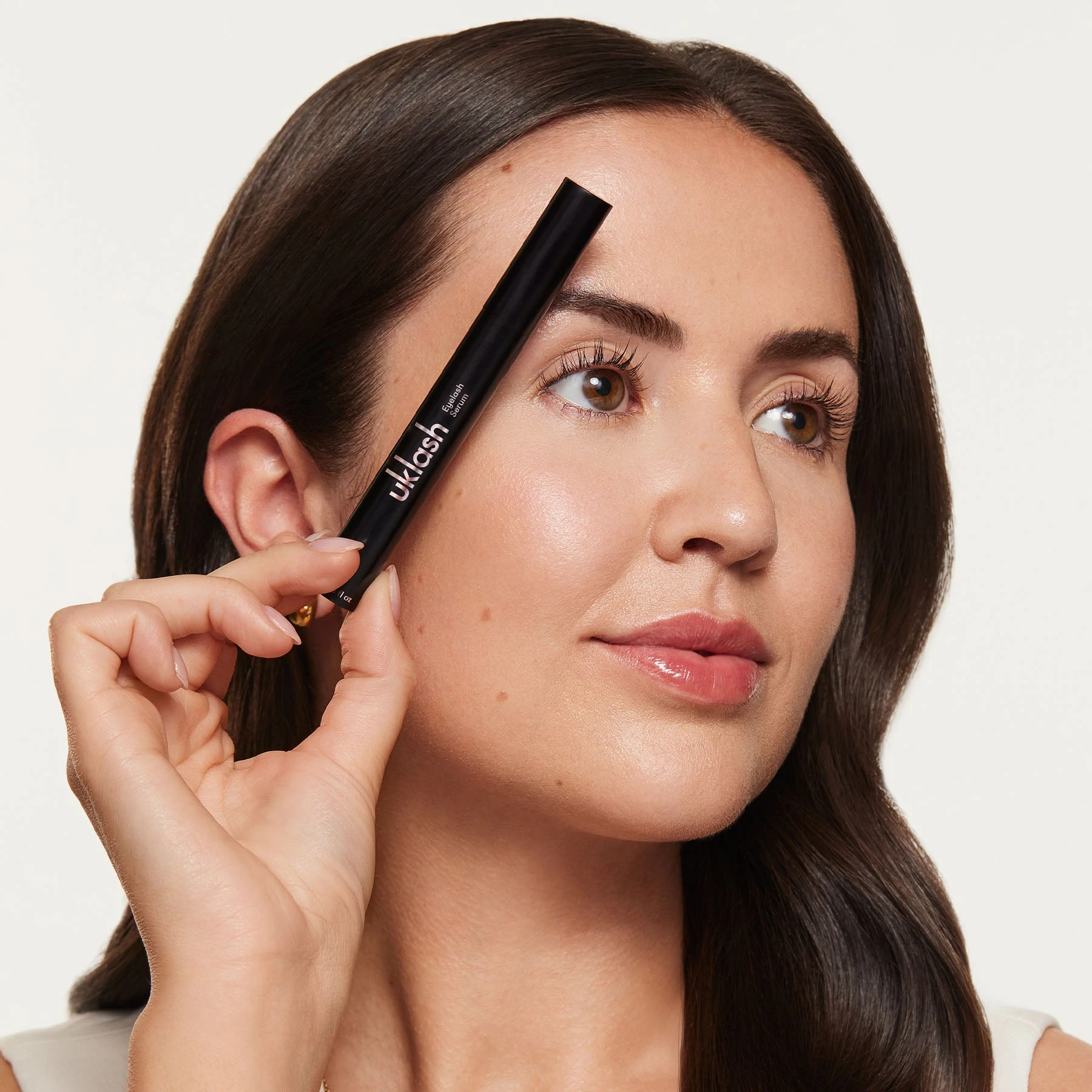 A woman with long eyelashes holds a black UKLASH Eyelash Serum tube near her right eye.