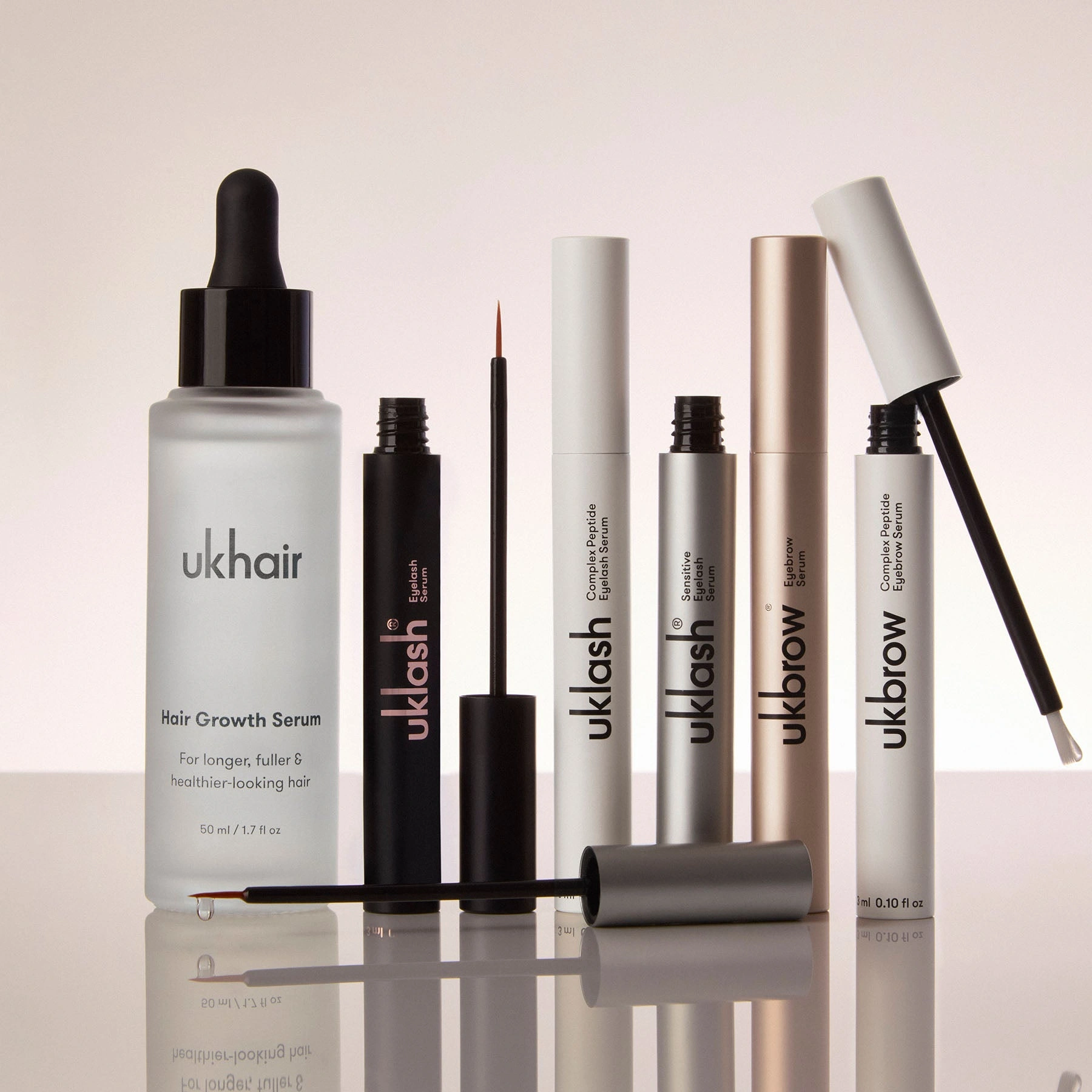 A lineup of haircare and beauty serums is displayed, including UKHAIR Hair Growth Serum, three UKLASH Eyelash Serums, and two UKBROW Eyebrow Serums. Several bottles are open, showing the dropper and brush applicators.