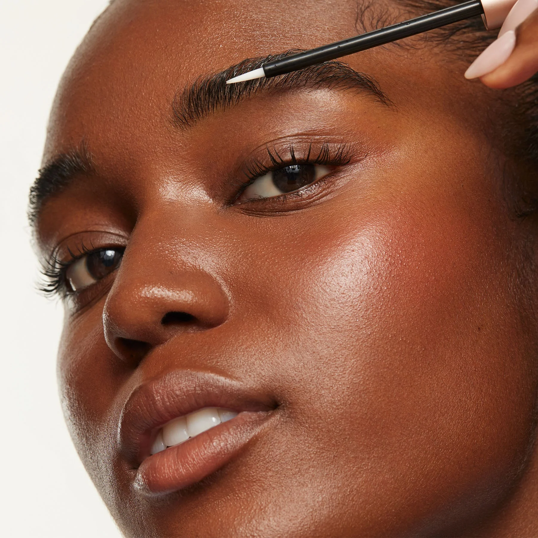 A woman with glowing skin applies UKBROW serum to their eyebrow with an applicator wand.